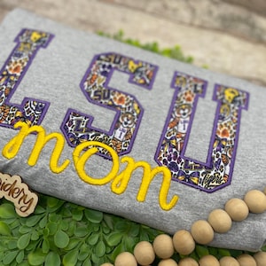 May include: A heather grey t-shirt featuring the letters LSU in a purple-bordered applique with a patterned print of tigers, paw prints, and bows. Below the letters, the word mom is embroidered in a bright yellow cursive script font.