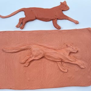 Può includere: Due sculture in terracotta di un gatto che corre. Una scultura è rialzata rispetto all'altra, che è incassata in una placca rettangolare in terracotta.