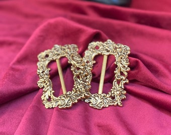 May include: Two ornate gold-toned metal buckles with intricate floral designs. The buckles are on a deep red velvet surface.