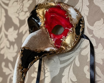 Può includere: Una maschera veneziana da carnevale bianca, nera e dorata con un lungo naso curvo. La maschera ha un foro per gli occhi rosso bordato d'oro ed è decorata con dettagli intricati.