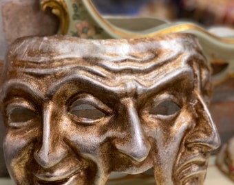 Può includere: Una maschera teatrale dipinta in oro e argento con due volti, uno sorridente e l'altro accigliato. La maschera è in legno e ha una superficie testurizzata.