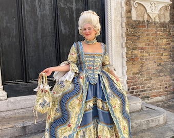 May include: A woman in a blue and gold patterned dress with a white wig and a small blue bag. She is standing on a set of stone steps in front of a building.