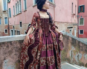 May include: A woman in a burgundy and floral patterned dress with a velvet trim. She is wearing a white lace collar and has her hair styled in a large, elaborate updo with a pink flower.