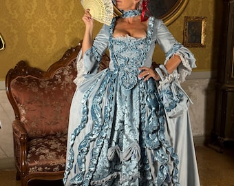 Peut inclure: Une robe bleu clair de style rococo avec des détails complexes, notamment des volants et des nœuds. La robe a des manches longues et un sac assorti. La femme porte une perruque blanche et tient un éventail. Le fond comprend des meubles et des œuvres d'art anciens.