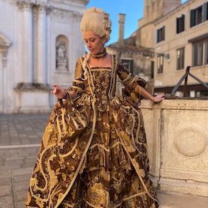 May include: A woman in a brown and gold brocade gown with a white wig and a gold necklace. The gown has a fitted bodice and a full skirt with ruffles. The woman is standing in front of a stone building.