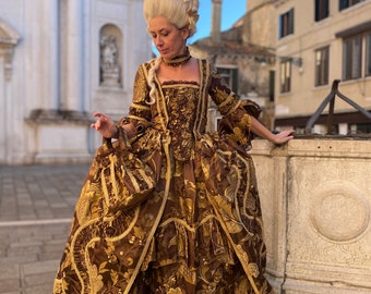 May include: A woman in a brown and gold brocade gown with a white wig and a gold necklace. The gown has a fitted bodice and a full skirt with ruffles. The woman is standing in front of a stone building.