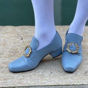 Puede incluir: Un par de zapatos azul claro con hebillas doradas. Los zapatos tienen un tac&oacute;n bajo y una punta puntiaguda.