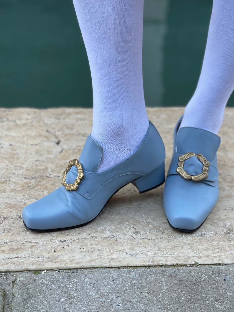 Puede incluir: Un par de zapatos azul claro con hebillas doradas. Los zapatos tienen un tac&oacute;n bajo y una punta puntiaguda.
