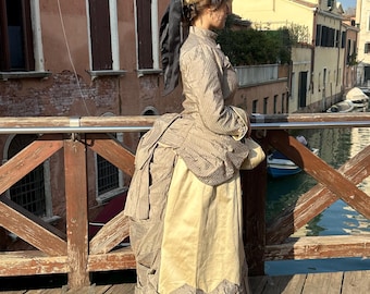 May include: A woman wearing a brown and white striped dress with a large bustle. She is standing on a wooden bridge over a canal. She is wearing a large hat with feathers and a black ribbon.