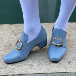 Puede incluir: Un par de zapatos de tac&oacute;n azul claro con hebillas doradas. Los zapatos tienen una punta cuadrada y un tac&oacute;n bajo.