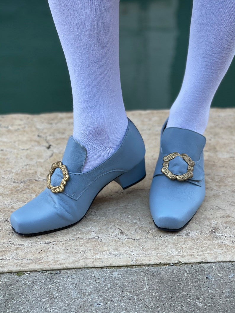 Puede incluir: Un par de zapatos de tac&oacute;n azul claro con hebillas doradas. Los zapatos tienen una punta cuadrada y un tac&oacute;n bajo.