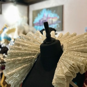 May include: A cream-colored, ruffled Elizabethan collar on a black mannequin. The collar features a detailed, repeating floral pattern and is displayed in a shop setting. The ruff is a historical costume element, often associated with Renaissance and Elizabethan fashion.