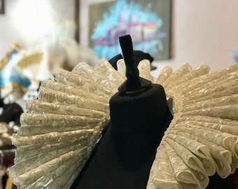 May include: A cream-colored, ruffled Elizabethan collar on a black mannequin. The collar features a detailed, repeating floral pattern and is displayed in a shop setting. The ruff is a historical costume element, often associated with Renaissance and Elizabethan fashion.