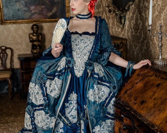 Könnte beinhalten: Eine Frau in einem historischen Kostüm, trägt ein blaues und silbernes Kleid mit aufwendigen Details. Das Kleid hat einen tiefen Ausschnitt, gerüschte Ärmel und einen weiten Rock. Sie trägt eine graue Perücke mit roten Blumen und ein Halsband. Sie hält einen Fächer.