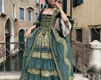 May include: A woman in a green and gold historical gown stands on a bridge in Venice. The dress features a fitted bodice, a full skirt with ruffles, and gold trim. The sleeves have lace cuffs, and the woman wears a matching wig and shoes.