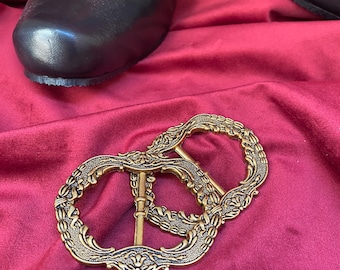 May include: Two antique gold-colored metal shoe buckles with ornate floral designs rest on a deep red velvet surface. Black leather shoes with matching gold buckles are in the background, suggesting a historical or costume theme.
