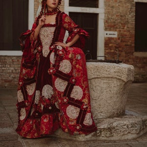 May include: A woman in a red and white patterned dress with a white lace overlay and a red velvet sash. She is wearing a white wig with a red ribbon and is sitting on a stone well.