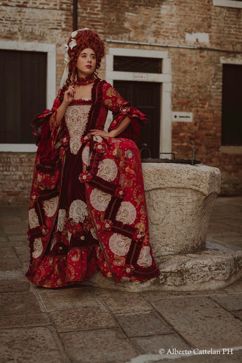 May include: A woman in a red and white patterned dress with a white lace overlay and a red velvet sash. She is wearing a white wig with a red ribbon and is sitting on a stone well.