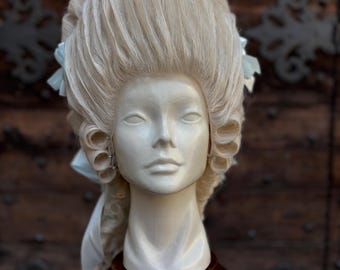 May include: A light blonde wig styled in the Rococo fashion, featuring high volume and curls. The wig is displayed on a white mannequin head atop a dark wooden stand. Light blue ribbons accent the hairstyle, set against a dark wood background.