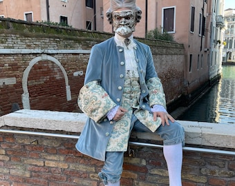 May include: A person wearing a blue velvet jacket with a white and gold patterned waistcoat and white knee-high socks. They are wearing a silver mask with a white wig and sitting on a brick wall.