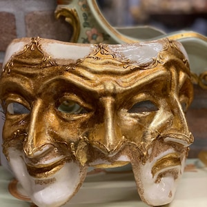 Może przedstawiać: Złota i biała wenecka maska z trzema twarzami, każda z innym wyrażeniem. Maska jest wykonana z papier-mâché i ozdobiona złotem.