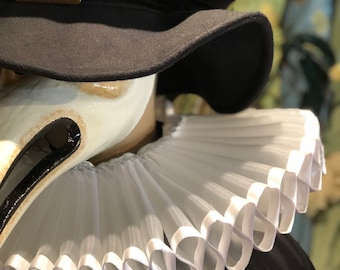 May include: A white ruffled collar with a white satin trim. The collar is worn over a black garment. A black hat with a gold buckle is worn on top of the collar.
