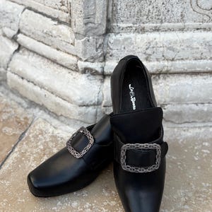 May include: A pair of black loafers featuring a square toe and a prominent rectangular buckle with an intricate silver-toned filigree pattern. The interior insole is printed with the brand name La Bauta in white script lettering.