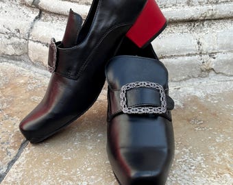 May include: A pair of black leather shoes with a square toe and a block heel in a contrasting red color. Each shoe features a decorative silver-toned rectangular buckle with an intricate textured pattern across the front strap.