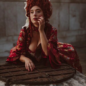 May include: A woman in a red and gold brocade dress with a white lace collar and a large red wig with white ribbons. She is sitting on a stone well with a wooden lid.
