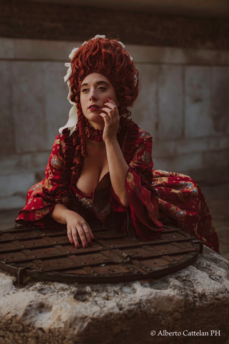 May include: A woman in a red and gold brocade dress with a white lace collar and a large red wig with white ribbons. She is sitting on a stone well with a wooden lid.