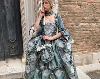 May include: A woman wearing a blue and silver gown with a white lace overlay and a large white wig. She is standing in front of a brick wall.
