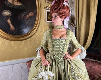 May include: A woman in a green and gold brocade gown with a large, elaborate wig. She is standing in a room with a painting on the wall behind her.