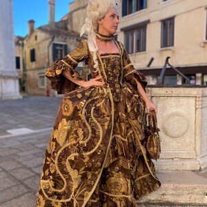 May include: A woman in a brown and gold brocade gown with a white wig and a small handbag. The gown has a floral pattern and gold trim. The woman is standing on a stone step.