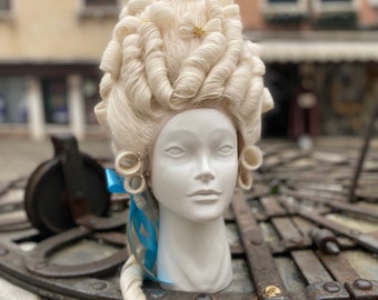 May include: A white wig with large, curled sections and gold accents. The wig is styled in a traditional, elaborate fashion. The wig is displayed on a white mannequin head.