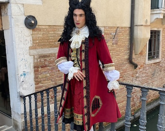 May include: A person wearing a red and gold brocade coat with a white lace collar and cuffs. The coat has a long, flowing skirt and is trimmed with black braid. The person is wearing a white wig with a black bow on top. The person is standing on a stone staircase with a railing.