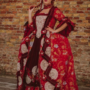 May include: A woman in a red and white dress with a large, elaborate skirt and a white lace bodice. She is wearing a white lace headpiece and has red hair. The dress is decorated with floral patterns and lace trim. The woman is standing in front of a brick wall.