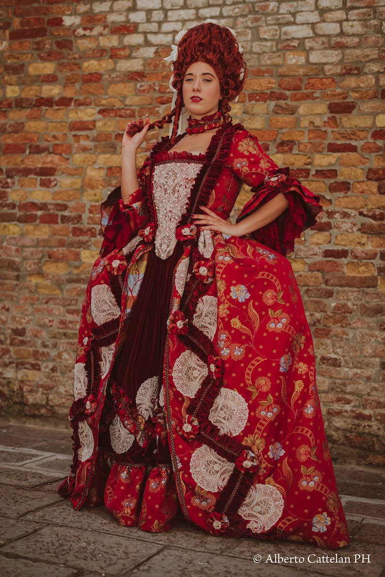 May include: A woman in a red and white dress with a large, elaborate skirt and a white lace bodice. She is wearing a white lace headpiece and has red hair. The dress is decorated with floral patterns and lace trim. The woman is standing in front of a brick wall.