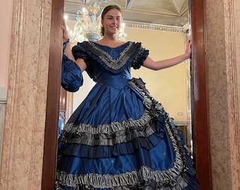 May include: A woman in a blue and black ball gown with a ruffled skirt stands in a doorway. The gown has a fitted bodice and a full skirt with multiple layers of ruffles. The woman is smiling and looking at the camera.