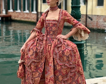 May include: A woman wearing a red and gold brocade gown with a white wig and a lace collar. The gown has a fitted bodice and a full skirt with ruffles. She is standing next to a green lamppost.