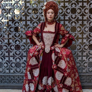 May include: A woman in a red and gold brocade dress with white lace trim stands in front of a metal gate. The dress has a fitted bodice and a full skirt. The woman has red hair and is wearing a pearl necklace.