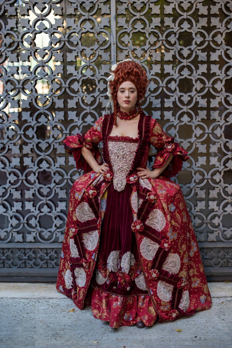May include: A woman in a red and gold brocade dress with white lace trim stands in front of a metal gate. The dress has a fitted bodice and a full skirt. The woman has red hair and is wearing a pearl necklace.