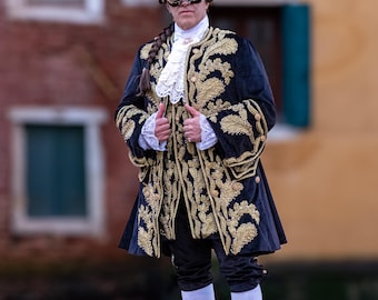May include: A person wearing a black velvet jacket with gold embroidery, a white lace shirt, and a black mask. They are wearing white socks and black shoes with gold buckles. The person is standing in front of a building with a window.