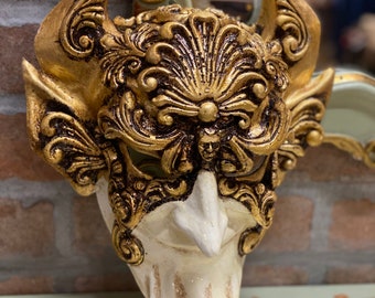 May include: A white and gold Venetian masquerade mask with intricate details and a long, pointed nose.