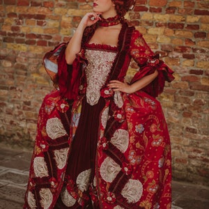 May include: A woman in a red and gold brocade dress with white lace accents. The dress has a fitted bodice and a full skirt. She is wearing a white wig with flowers and a red choker necklace. The background is a brick wall.