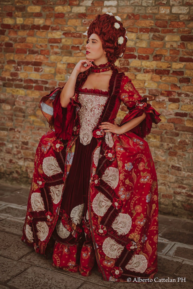 May include: A woman in a red and gold brocade dress with white lace accents. The dress has a fitted bodice and a full skirt. She is wearing a white wig with flowers and a red choker necklace. The background is a brick wall.