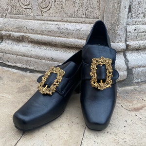 May include: A pair of black leather shoes with gold ornate buckles. The shoes have a square toe and a low heel.