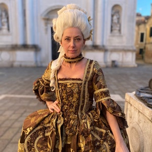 May include: A woman wearing a brown and gold brocade dress with a white wig and a gold choker necklace. The dress has a fitted bodice and a full skirt. The woman is standing in front of a white building.