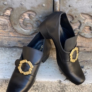 May include: A pair of black leather shoes with gold buckles. The shoes have a pointed toe and a low heel.