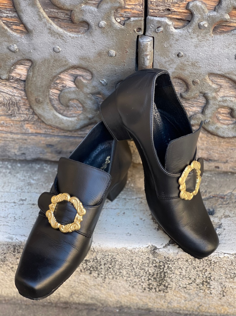 May include: A pair of black leather shoes with gold buckles. The shoes have a pointed toe and a low heel.