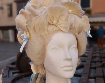 May include: A light blonde wig styled in a historical rococo fashion, featuring elaborate curls and floral accents. The wig is adorned with small pearls and light blue ribbons, displayed on a white mannequin head.